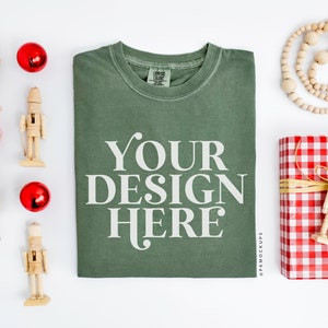 May include: A green t-shirt with the text "YOUR DESIGN HERE" printed on the front. The t-shirt is surrounded by Christmas decorations, including nutcrackers, ornaments, and a gift wrapped in red and white gingham.