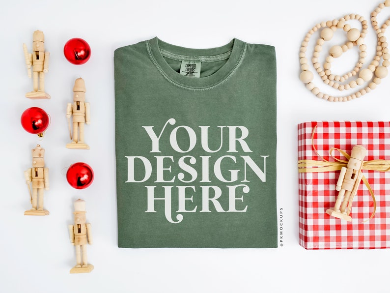May include: A green t-shirt with the text "YOUR DESIGN HERE" printed on the front. The t-shirt is surrounded by Christmas decorations, including nutcrackers, ornaments, and a gift wrapped in red and white gingham.