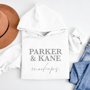 May include: White hooded sweatshirt with the text "PARKER & KANE mockups" on the front. The sweatshirt is laid flat on a white surface with a brown hat and blue jeans.