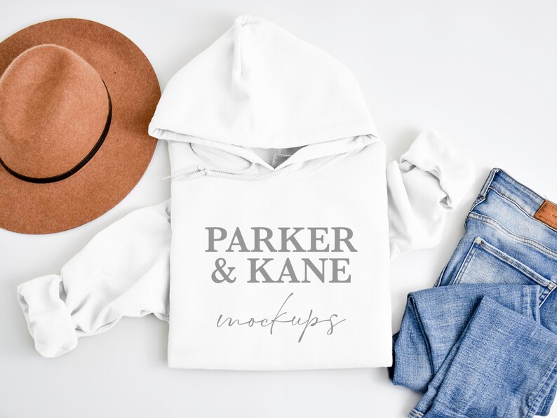 May include: White hooded sweatshirt with the text "PARKER & KANE mockups" on the front. The sweatshirt is laid flat on a white surface with a brown hat and blue jeans.