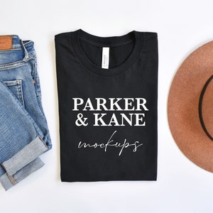 May include: A black t-shirt with the text "PARKER & KANE mockups" in white. The t-shirt is folded and laid flat on a white surface. There are blue jeans and a brown hat beside the t-shirt.