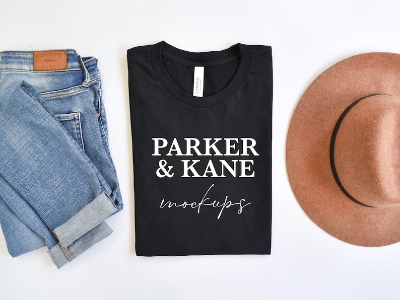 May include: A black t-shirt with the text "PARKER & KANE mockups" in white. The t-shirt is folded and laid flat on a white surface. There are blue jeans and a brown hat beside the t-shirt.