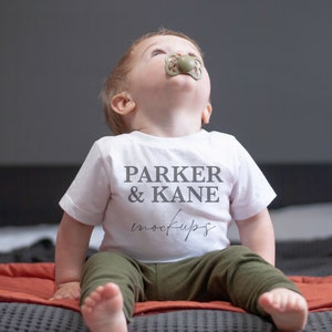 Op de afbeelding: Een witte baby romper met de tekst "PARKER & KANE mockups" op de voorkant. Een baby draagt de romper en zit op een bed met een rode deken.