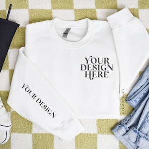 May include: A white crewneck sweatshirt with the text "YOUR DESIGN HERE" and "YOUR DESIGN" printed in black. The image also includes a black tumbler with a straw, white sneakers, and blue jeans, all arranged on a green and white checkered surface.