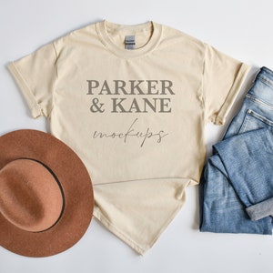 May include: A beige t-shirt with the text "Parker & Kane mockups" printed on the front. The t-shirt is laid flat on a white surface with a brown hat and blue jeans.