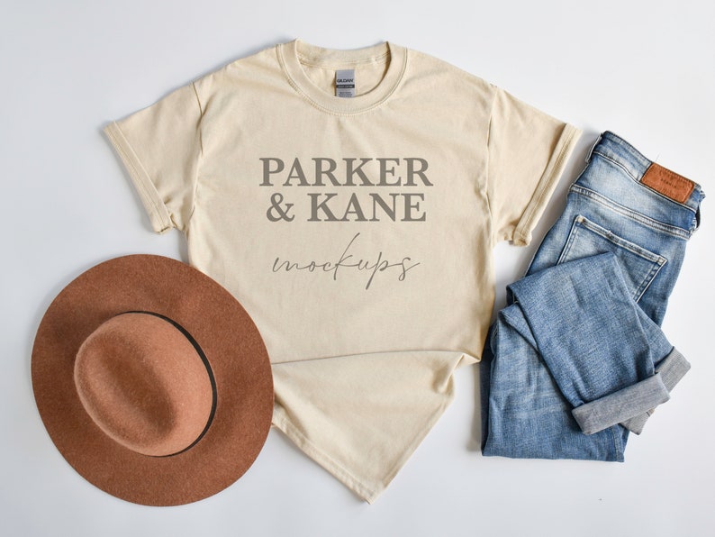 May include: A beige t-shirt with the text "Parker & Kane mockups" printed on the front. The t-shirt is laid flat on a white surface with a brown hat and blue jeans.