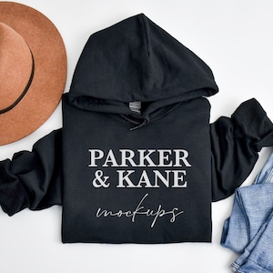 Op de afbeelding: Zwart hoodie met de tekst "PARKER & KANE mockups" bedrukt op de voorkant. De hoodie ligt plat op een wit oppervlak met een bruine hoed en een blauwe jeans.