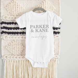 May include: White baby onesie with short sleeves, hanging on a gray hanger. The onesie has the text "PARKER & KANE mockups" printed on the front. The background features a woven wall hanging with cream and black accents.