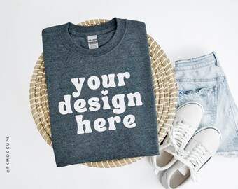 May include: A dark gray t-shirt with the text "your design here" in white. The t-shirt is laying on a woven basket with a pair of white sneakers and light blue denim shorts beside it.