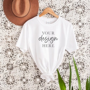 Op de afbeelding: Wit T-shirt met korte mouwen hangend aan een houten hanger tegen een witte en zwarte gepatroneerde achtergrond. Het shirt heeft de tekst "YOUR design HERE" op de voorkant.
