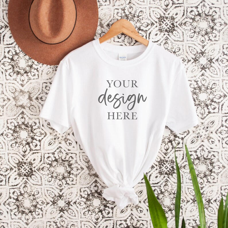 Op de afbeelding: Wit T-shirt met korte mouwen hangend aan een houten hanger tegen een witte en zwarte gepatroneerde achtergrond. Het shirt heeft de tekst "YOUR design HERE" op de voorkant.