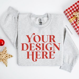 May include: A light gray sweatshirt with the text "YOUR DESIGN HERE" in red. The sweatshirt is surrounded by Christmas decorations, including gold stars, red and white ornaments, and a wrapped gift.
