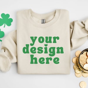 Könnte beinhalten: Ein beiger Pullover mit dem Text "your design here" in grünen Buchstaben. Der Pullover ist gefaltet und liegt flach auf einer weißen Oberfläche. Im Hintergrund befinden sich grüne Kleeblatt-Ausschnitte und eine Schale mit Goldmünzen.