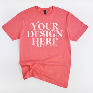 May include: A coral colored t-shirt with the text "YOUR DESIGN HERE" in white. The text is centered on the shirt and is surrounded by a white border.