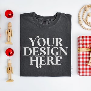May include: A flat lay mockup featuring a dark gray t-shirt with the text "YOUR DESIGN HERE" printed on the front. The t-shirt is surrounded by red ornaments, wooden nutcrackers, a wooden bead garland, and a red and white gingham gift box with a wooden nutcracker on top.