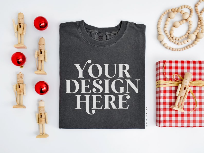 May include: A flat lay mockup featuring a dark gray t-shirt with the text "YOUR DESIGN HERE" printed on the front. The t-shirt is surrounded by red ornaments, wooden nutcrackers, a wooden bead garland, and a red and white gingham gift box with a wooden nutcracker on top.