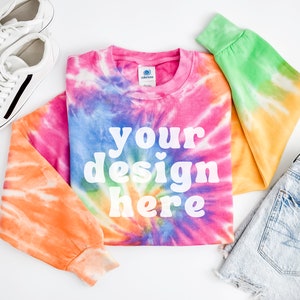 Może przedstawiać: Dług rękawowa bluza w stylu tie-dye z napisem "your design here" w kolorze białym. Bluza leży płasko na białym tle, obok niej znajdują się białe buty sportowe i jasnoniebieskie dżinsowe spodenki.