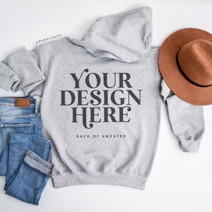 May include: A heather gray hoodie with the text "YOUR DESIGN HERE" and the words "BACK OF SWEATER" below it. A brown hat and blue jeans are next to the hoodie. The hoodie and jeans are folded. The hat is brown with a black band.
