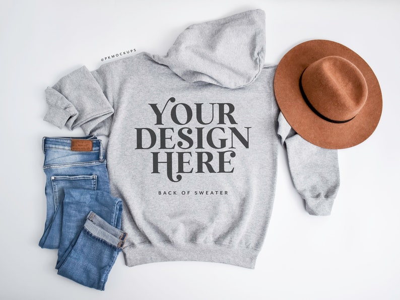 May include: A heather gray hoodie with the text "YOUR DESIGN HERE" and the words "BACK OF SWEATER" below it. A brown hat and blue jeans are next to the hoodie. The hoodie and jeans are folded. The hat is brown with a black band.