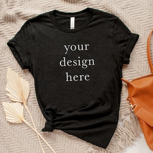 以下が含まれることがあります： 前面に「your design here」という文字が印刷された黒いTシャツ。Tシャツは、ベージュのテクスチャのある表面に、茶色の革のバッグと乾燥したヤシの葉と一緒に置かれています。