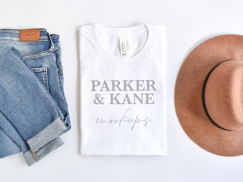 May include: White t-shirt with the text "Parker & Kane mockups" printed in gray. The t-shirt is laid flat on a white surface next to a pair of blue jeans and a brown fedora.