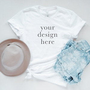 May include: A white short-sleeved t-shirt with the text your design here printed in black on the front, paired with a light brown wide-brimmed hat and light blue denim shorts featuring a white floral pattern and a frayed hemline.