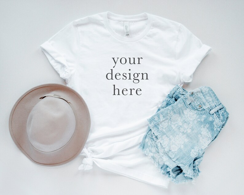 May include: A white short-sleeved t-shirt with the text your design here printed in black on the front, paired with a light brown wide-brimmed hat and light blue denim shorts featuring a white floral pattern and a frayed hemline.