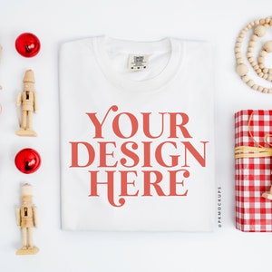 May include: White t-shirt with the text "YOUR DESIGN HERE" in red on a white background. The t-shirt is surrounded by red ornaments, wooden nutcrackers, and a red and white checkered gift box with a wooden nutcracker on top.