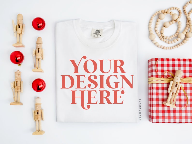 May include: White t-shirt with the text "YOUR DESIGN HERE" in red on a white background. The t-shirt is surrounded by red ornaments, wooden nutcrackers, and a red and white checkered gift box with a wooden nutcracker on top.