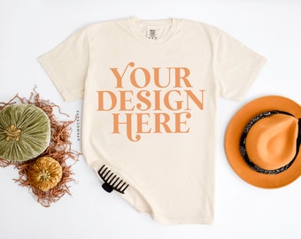 May include: Cream-colored t-shirt with the text "YOUR DESIGN HERE" in orange. The shirt is next to an orange hat with a black bandana, a black hair clip, and decorative pumpkins. The image is on a white background.