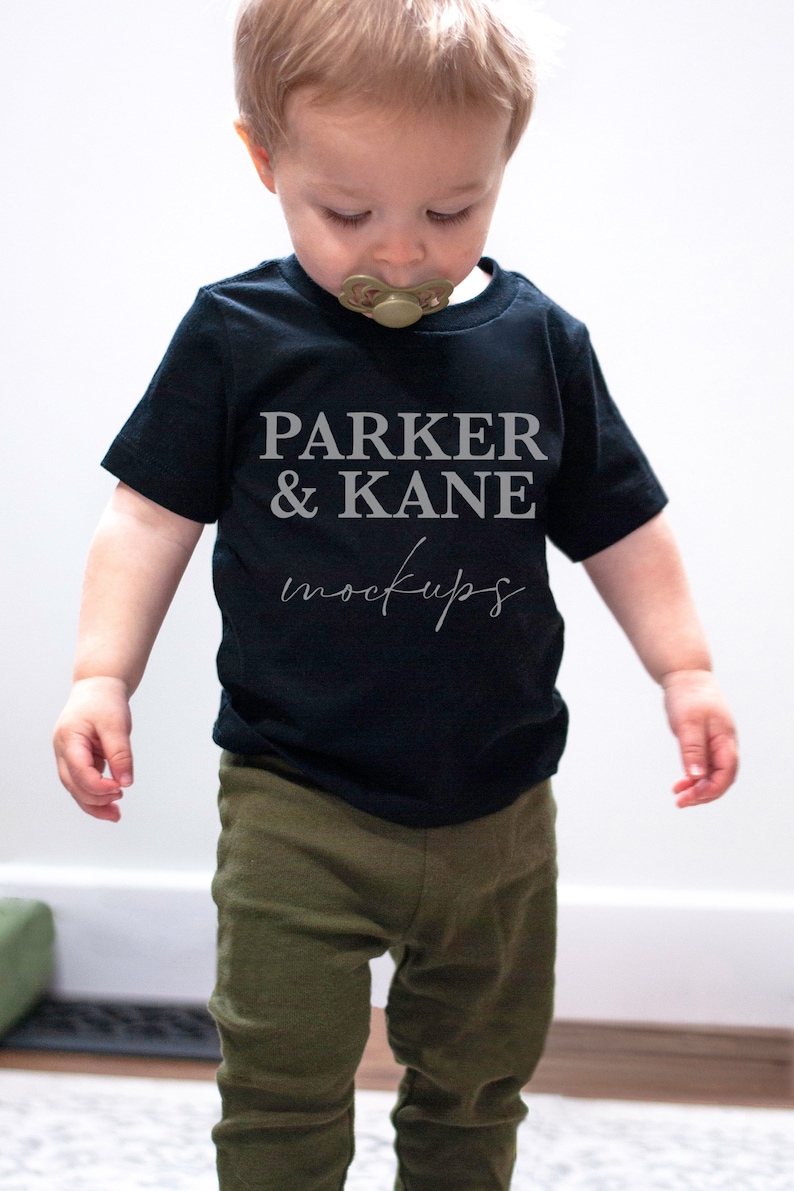 Pu&ograve; includere: Un bambino piccolo che indossa una maglietta nera con il testo "PARKER & KANE mockups" in lettere grigie. Il bambino indossa pantaloni verde oliva e guarda in basso.