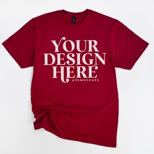 May include: A maroon t-shirt with short sleeves and a crew neck. The shirt has the text "YOUR DESIGN HERE" in white on the front. The text is in a serif font. The shirt is laid flat on a white background.