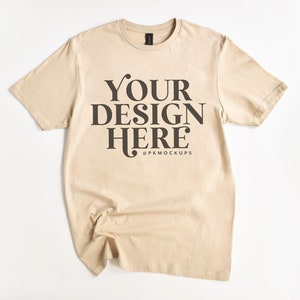 May include: A beige t-shirt with short sleeves and a crew neck. The shirt has the text "YOUR DESIGN HERE" printed on the front in black.