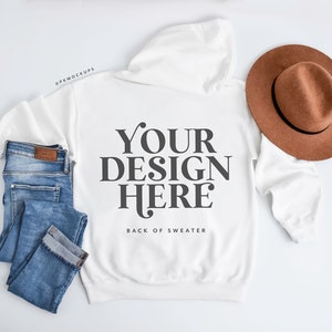 May include: A white hoodie with the text "YOUR DESIGN HERE" on the back, paired with blue jeans and a brown hat. The hoodie is laid flat, showcasing the design. The jeans are folded neatly, and the hat is placed on the side.