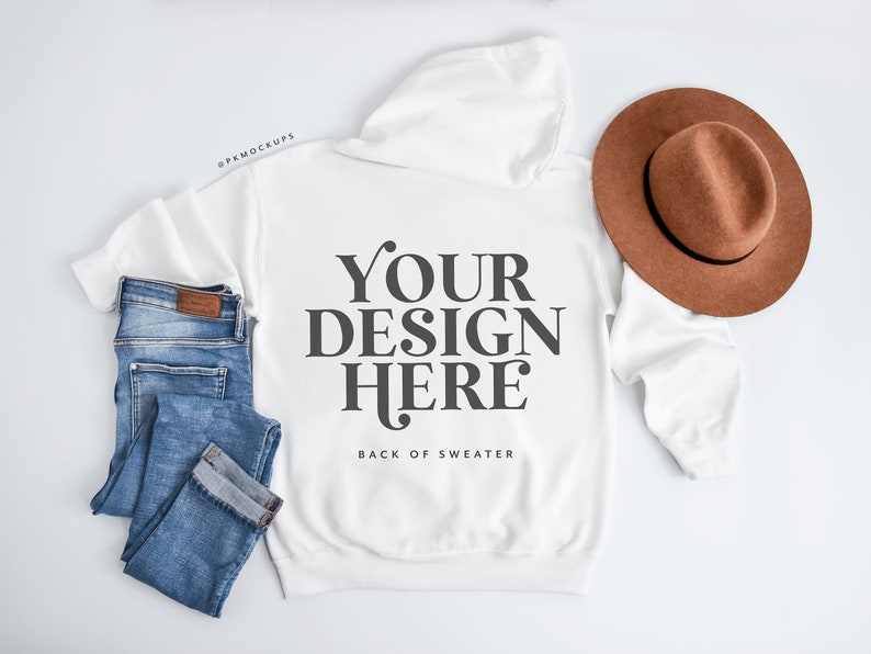 May include: A white hoodie with the text "YOUR DESIGN HERE" on the back, paired with blue jeans and a brown hat. The hoodie is laid flat, showcasing the design. The jeans are folded neatly, and the hat is placed on the side.