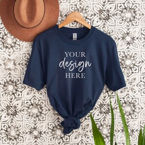 Op de afbeelding: Een marineblauw t-shirt met korte mouwen hangt aan een houten hanger tegen een witte en zwarte gepatroneerde achtergrond. Het shirt heeft de tekst "YOUR design HERE" op de voorkant.