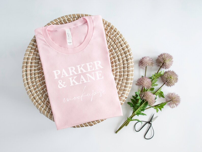May include: A pink t-shirt with the text "PARKER & KANE mockups" printed on it. The t-shirt is folded and placed on a woven basket.
