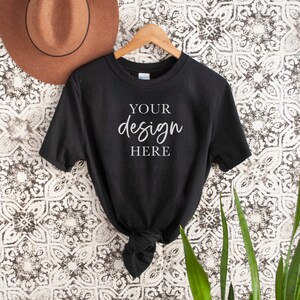 Op de afbeelding: Zwart T-shirt met korte mouwen hangend aan een houten hanger. Het shirt heeft de tekst "YOUR design HERE" in wit op de voorkant.