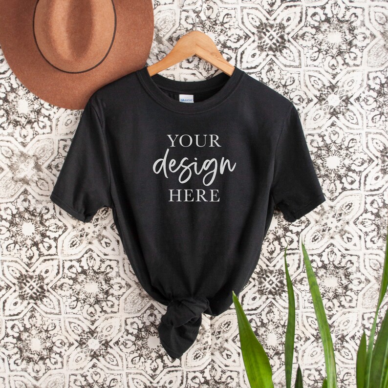 Op de afbeelding: Zwart T-shirt met korte mouwen hangend aan een houten hanger. Het shirt heeft de tekst "YOUR design HERE" in wit op de voorkant.