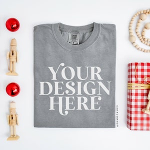 May include: A gray t-shirt with the text "YOUR DESIGN HERE" printed on the front. The t-shirt is folded and laid flat on a white surface. There are several red ornaments and wooden nutcracker figurines around the t-shirt.