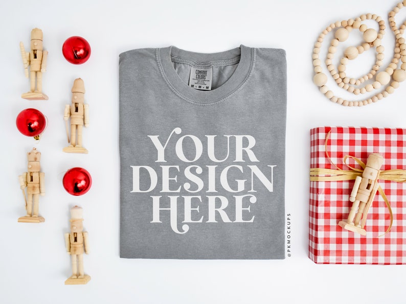 May include: A gray t-shirt with the text "YOUR DESIGN HERE" printed on the front. The t-shirt is folded and laid flat on a white surface. There are several red ornaments and wooden nutcracker figurines around the t-shirt.