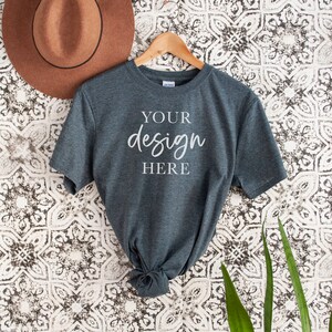Op de afbeelding: Een donkergrijs t-shirt met korte mouwen hangt aan een houten hanger tegen een witte en zwarte gepatroneerde achtergrond. Het shirt heeft de tekst "YOUR design HERE" op de voorkant.