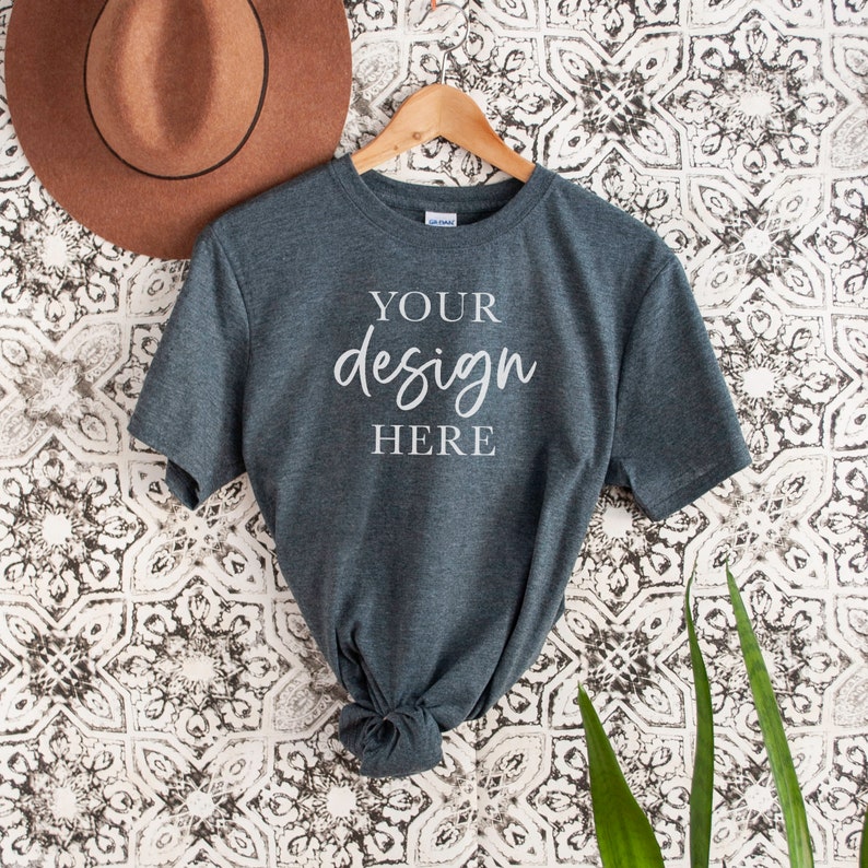Op de afbeelding: Een donkergrijs t-shirt met korte mouwen hangt aan een houten hanger tegen een witte en zwarte gepatroneerde achtergrond. Het shirt heeft de tekst "YOUR design HERE" op de voorkant.