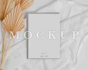 May include: A vertical rectangular greeting card mockup with a light gray border, featuring the text MOCKUP in large white letters and 5 x 7 jpg + psd printed in smaller text below. The card is positioned on a white fabric surface.