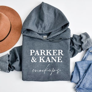 May include: A grey hooded sweatshirt with the text "PARKER & KANE mockups" printed on the front. The sweatshirt is laid flat on a white surface with a brown hat and blue jeans.
