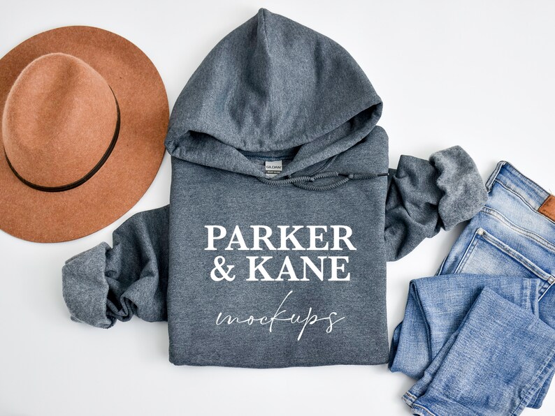 May include: A grey hooded sweatshirt with the text "PARKER & KANE mockups" printed on the front. The sweatshirt is laid flat on a white surface with a brown hat and blue jeans.