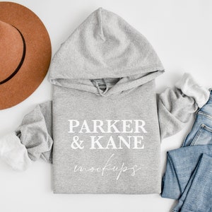 May include: A grey hooded sweatshirt with the text "PARKER & KANE mockups" printed on the front. The sweatshirt is laid flat on a white surface with a brown hat and blue jeans.