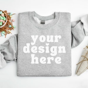 May include: A heather gray crewneck sweatshirt with the text "your design here" in white. The image includes Easter-themed decorations such as a bowl of pastel-colored eggs and a carrot-shaped decoration.