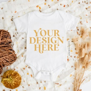May include: White baby onesie with the text "YOUR DESIGN HERE" in gold on a white background. The onesie is lying on a white and brown patterned fabric with dried flowers and a small pumpkin.