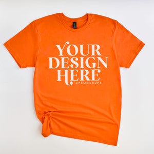 May include: Orange t-shirt with the text "YOUR DESIGN HERE" in white lettering. The shirt is crumpled and laid flat on a white surface.